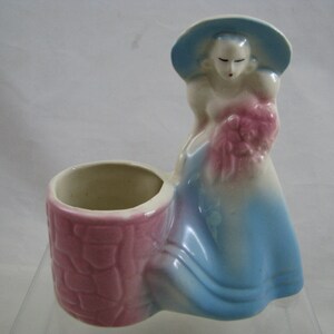 May include: A vintage ceramic planter featuring a woman in a blue dress and hat holding a bouquet of pink flowers. The planter has a pink and white glazed finish and a separate planter pot.