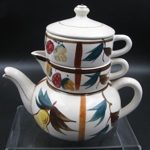 May include: A white ceramic tea set with a teapot and two stacked teacups. The set is decorated with brown vertical stripes, green leaves, and colorful fruit designs. The teapot has a curved spout and handle.