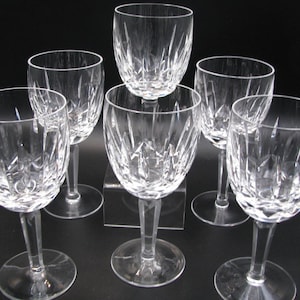 May include: Six clear crystal wine glasses with a cut glass design. The glasses have a rounded bowl shape, a long stem, and a circular base. The glasses are arranged on a dark surface, showcasing their intricate patterns and clarity.