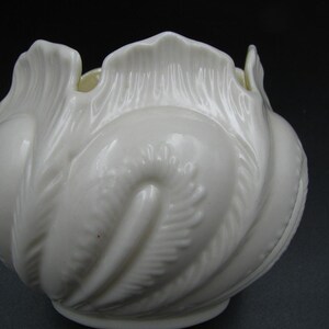 May include: A white ceramic vase with a ruffled, petal-like design. The vase has a glossy finish and a swirling, textured pattern. The opening is scalloped, and the overall shape is rounded.