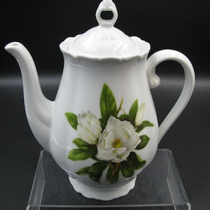 May include: A white porcelain teapot featuring a scalloped rim and a matching lid with a decorative finial. The body is adorned with a detailed botanical illustration of white magnolia flowers and green leaves centered on the front surface.