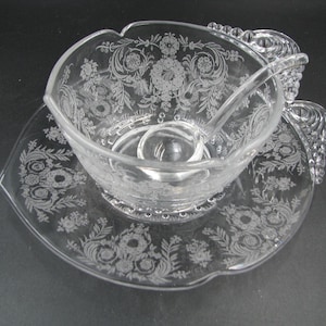 May include: A clear glass punch bowl set featuring a delicate etched floral pattern across the surface. The set includes a matching glass ladle and a wide, leaf-shaped serving plate with a textured, beaded handle detail on the side.