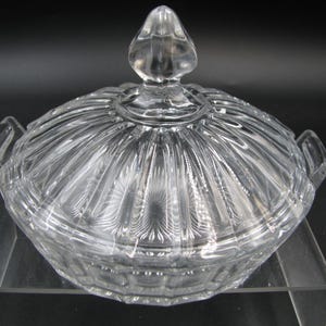May include: A clear glass serving dish with a matching lid, featuring a ribbed pattern and two side handles. The lid is topped with a pointed finial, and the base displays a pressed geometric design, creating a reflective and transparent appearance.