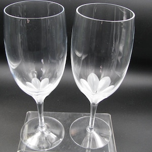May include: Two clear wine glasses with a white flower design etched at the base of the bowl. The glasses have a slender stem and a round base. The background is dark, highlighting the transparency of the glass.