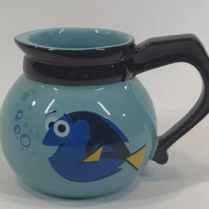 May include: A light blue ceramic coffee mug featuring a cartoon image of Dory from Finding Nemo. The mug has a black handle and a black rim. Bubbles are depicted on the side of the mug.