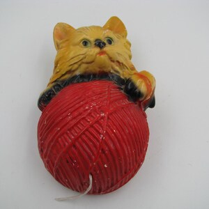 May include: A molded decorative figure of a yellow cat with black accents resting its paws on a large, round, textured ball of red yarn. The cat has green eyes and a small pink nose, while the red yarn features a visible white string end.