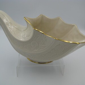 May include: A cream-colored ceramic cornucopia bowl featuring an embossed fruit motif on the exterior, a scalloped rim, and a gold-trimmed edge. The vessel has a curved, horn-shaped handle and rests on a circular base with a gold-colored trim.