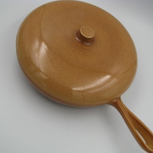 May include: A tan ceramic skillet with a lid and a handle. The skillet is oval-shaped and has a speckled texture. The lid has a small, round knob. The handle is curved and has an opening.