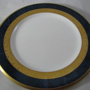 May include: A white ceramic dinner plate with a wide gold and blue rim. The rim features a marbled blue design with a gold border and intricate gold patterns.