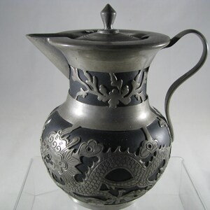 May include: A silver teapot with a black dragon design. The teapot has a lid and a handle. The dragon is embossed on the teapot.