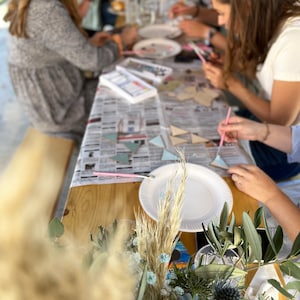 May include: A group of people are gathered around a table, painting wooden geometric shapes with colorful paints. The table is covered in a white tablecloth with a newspaper on top. There are paintbrushes, paint palettes, and other painting supplies on the table. The people are all wearing casual clothing and are having fun painting.