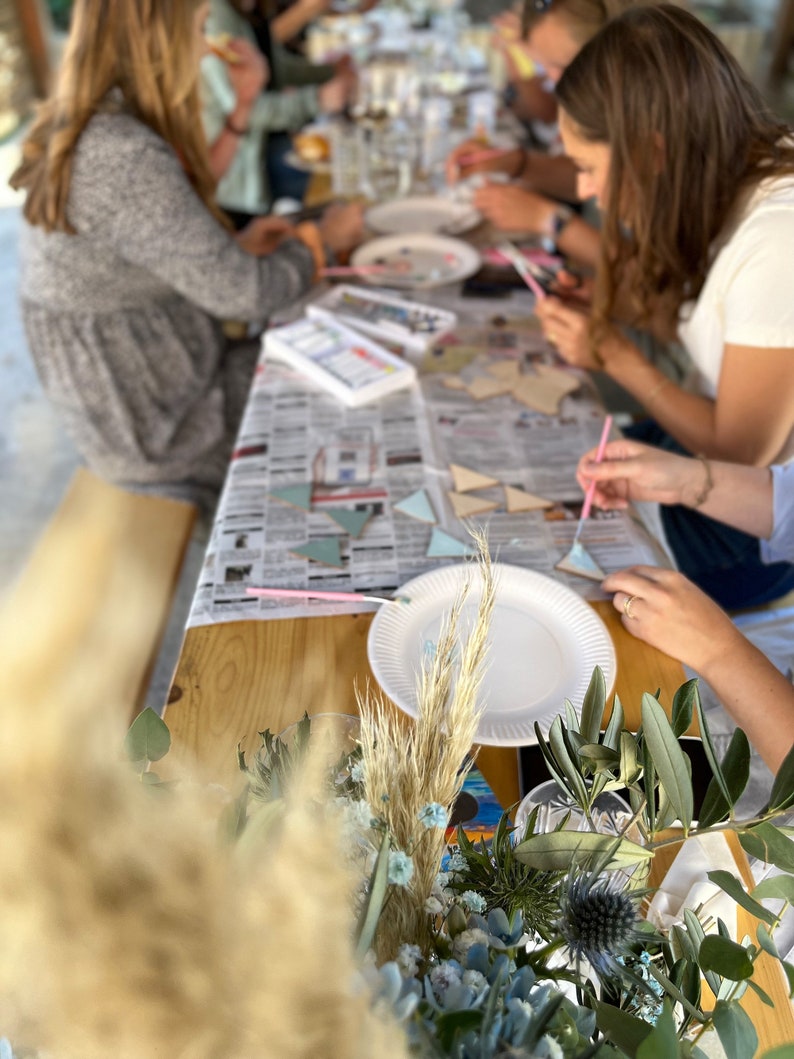 May include: A group of people are gathered around a table, painting wooden geometric shapes with colorful paints. The table is covered in a white tablecloth with a newspaper on top. There are paintbrushes, paint palettes, and other painting supplies on the table. The people are all wearing casual clothing and are having fun painting.