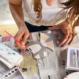 May include: A person paints a gray and white wooden animal cutout with a paintbrush. The cutout is shaped like a raccoon with a black nose and eyes. The person is using white paint to paint the animal's fur.