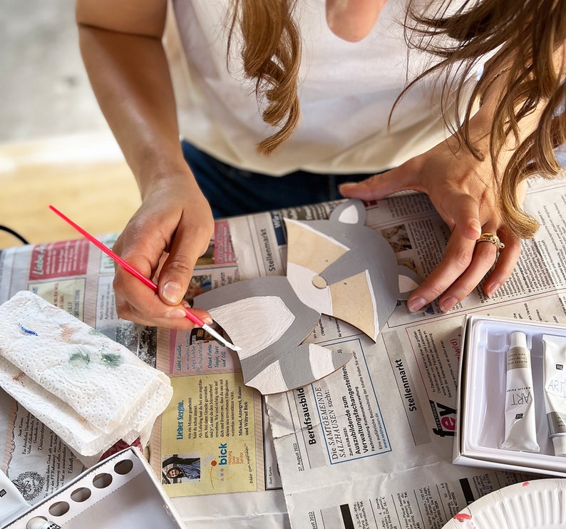 May include: A person paints a gray and white wooden animal cutout with a paintbrush. The cutout is shaped like a raccoon with a black nose and eyes. The person is using white paint to paint the animal's fur.