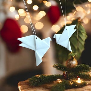 Op de afbeelding: Twee witte origami-duiven die aan een kerstboom hangen. De duiven zijn gemaakt van gevouwen papier en hebben een eenvoudig, elegant ontwerp.