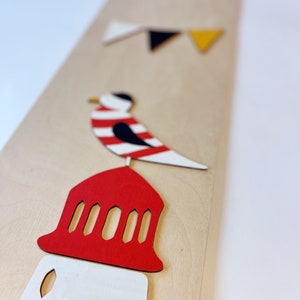 May include: A wooden wall decor featuring a red and white lighthouse, a black and white bird, and a string of colorful flags. The lighthouse has a red roof with white windows and a white base. The bird is perched on top of the lighthouse. The flags are red, black, white, and yellow.