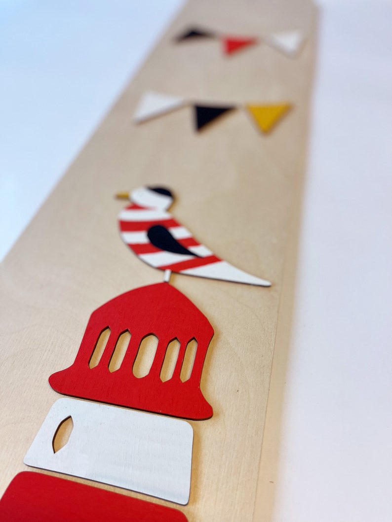 May include: A wooden wall decor featuring a red and white lighthouse, a black and white bird, and a string of colorful flags. The lighthouse has a red roof with white windows and a white base. The bird is perched on top of the lighthouse. The flags are red, black, white, and yellow.