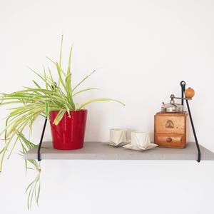 May include: A concrete shelf with black metal brackets holds a red pot with a green plant, a vintage coffee grinder, and two white ceramic cups.