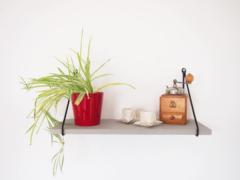May include: A concrete shelf with black metal brackets holds a red pot with a green plant, a vintage coffee grinder, and two white ceramic cups.