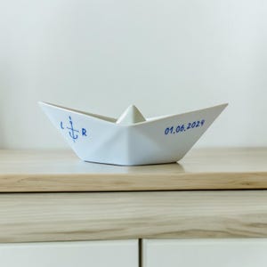 Op de afbeelding: Een wit keramieken papieren bootsculptuur met blauwe inktaccenten. De linkerkant toont een gestileerd ankersymbool geflankeerd door de letters L en R. De rechterkant bevat de datum 01.06.2024 gedrukt in een eenvoudig blauw schreefloos lettertype.
