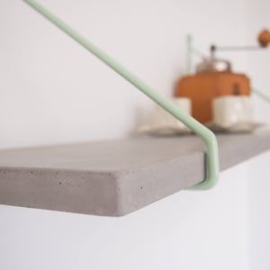 May include: A gray concrete shelf with light green metal brackets. The shelf is mounted to a white wall.
