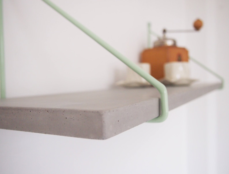 May include: A gray concrete shelf with light green metal brackets. The shelf is mounted to a white wall.