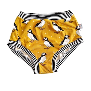 Puede incluir: Bragas amarillas mostaza con un estampado repetido de frailecillos en blanco y negro. La cintura y las aberturas de las piernas tienen ribetes a rayas blancas y negras. Pequeños triángulos blancos en el fondo.