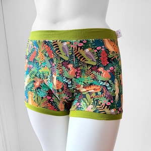 May include: Boxer briefs featuring a vibrant jungle print with tigers, monkeys, toucans, and parrots amidst lush green foliage on a dark background. The garment has a solid olive green elastic waistband and leg cuffs, with a small white tag on the side.