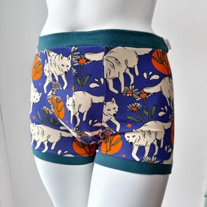 May include: Blue boxer shorts with a white wolf and floral pattern. The shorts have a teal waistband and trim. The design includes white wolves, orange circles, and white daisies on a blue background.