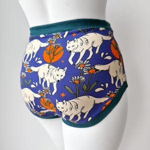 Puede incluir: Bragas de cintura alta con un fondo azul con un estampado repetido de lobos blancos, soles naranjas y margaritas blancas. La cintura y el ribete son de color verde azulado. La ropa interior se muestra en un maniquí blanco.