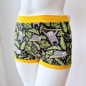 May include: Shorts featuring a black background with a repeating pattern of grey sloths hanging from and resting on tan branches, surrounded by green tropical leaves. The garment has a wide, solid mustard yellow waistband and leg cuffs.