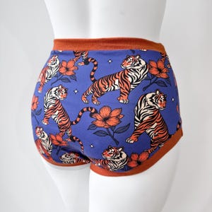 Könnte beinhalten: Hochtaillierte Unterwäsche mit blauem Hintergrund und einem sich wiederholenden Muster aus Tigern und orangefarbenen Blumen. Der Bund und die Zierleiste sind rostfarben. Die Tiger haben schwarz-orange Streifen.