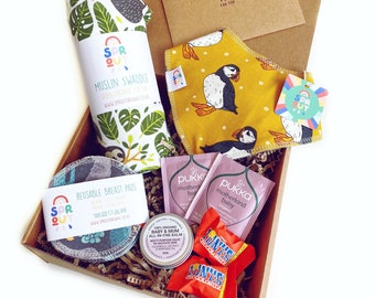 May include: A gift box filled with baby items, including a muslin swaddle with a sloth design, a bib with a puffin design, reusable breast pads, a baby balm, and two tea bags. The box is decorated with brown paper and has a small tag that says "A gift for you".