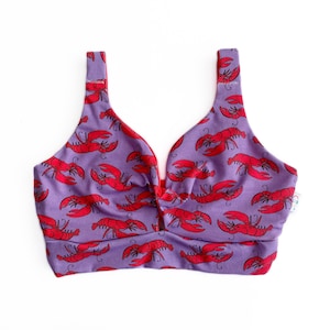 May include: A lavender-colored crop top with a repeating pattern of red lobsters. The top has a V-neck design and wide straps. The edges are trimmed in red.