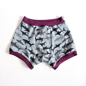 May include: Light blue boxer shorts with a repeating shark pattern in shades of gray and black. The waistband and leg openings are trimmed with a deep purple fabric. The shorts are made of a soft, comfortable material.