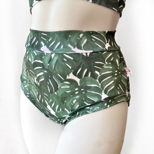 Peut inclure: Bas de maillot de bain taille haute avec un imprimé de feuilles tropicales dans des tons de vert et de blanc. Le tissu a un léger brillant et les bas ont une jambe haute.