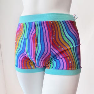 May include: Colorful boxer shorts with a rainbow wave pattern and small star accents. The waistband and leg openings are a solid turquoise color. The shorts are displayed on a white mannequin.