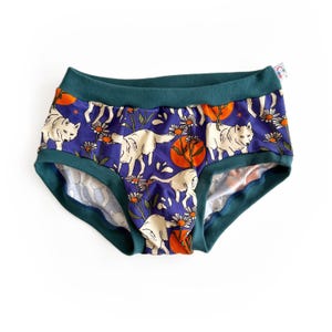 Peut inclure: Culotte or shorty avec un motif de loups blancs, de soleils oranges et de marguerites sur un fond bleu foncé. La culotte a une bordure et une ceinture vert d'eau. Une petite étiquette est attachée sur le côté.