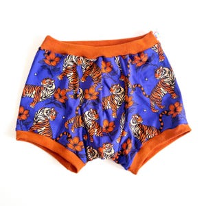 May include: Blue boxer shorts with an orange waistband and trim. The shorts feature a repeating pattern of orange tigers and flowers. The fabric is a vibrant blue, and the overall design is eye-catching and playful.