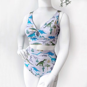 May include: White two-piece swimsuit with a dinosaur print. The top has a knotted front and a keyhole cutout. The bottoms are high-waisted.