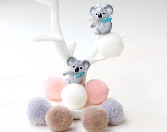 May include: Two gray koala bear charms with blue bows sitting on top of white and pink pom poms. The pom poms are on a white tree-shaped display stand.