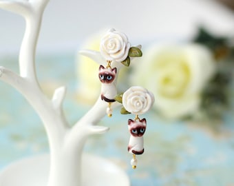 May include: A pair of dangle earrings featuring white porcelain roses and Siamese cats. The cats are sitting on the roses and have blue eyes. The earrings are hanging from a white tree branch.