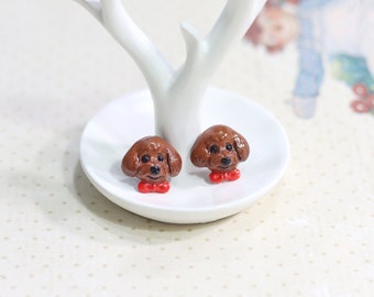 May include: A pair of brown poodle dog earrings with red bow ties. The earrings are displayed on a white ceramic tree stand.