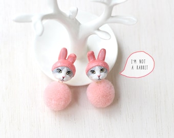 May include: A pair of pink pom-pom earrings with a cat wearing a pink bunny hat charm. The cat has gray fur and a white face. The earrings are on a white display stand. The text "I'm not a rabbit" is in a speech bubble.