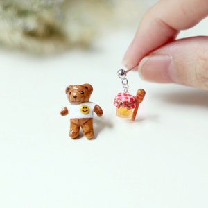 May include: A pair of handmade polymer clay earrings. One earring features a brown teddy bear wearing a white shirt with a yellow smiley face. The other earring features a small red and white checkered jar with a brown stick, representing honey.