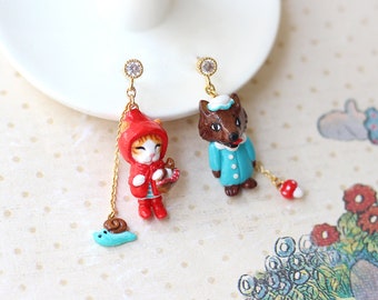 May include: A pair of dangle earrings featuring a red cat wearing a red hood and a blue wolf wearing a blue coat. The cat is holding a basket and the wolf is holding a red mushroom. The earrings are made of gold and have a clear crystal at the top.