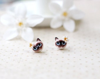 May include: A pair of small, white ceramic cat earrings with blue eyes and a white flower on top. The earrings are on a white background with a subtle polka dot pattern.