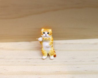 May include: A small ceramic figurine of a cat holding a skewer of dango, a Japanese sweet treat. The cat is orange and white with black eyes and a pink nose. The dango are white and round.