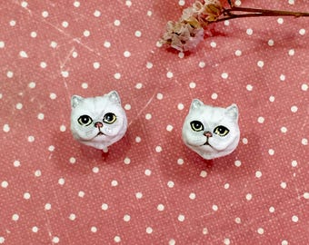 May include: A pair of white ceramic cat face earrings with yellow eyes. The earrings are on a pink background with white polka dots.