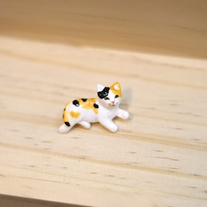 May include: A small ceramic figurine of a calico cat lying down. The cat is white with black and orange markings. The figurine is painted with realistic details.